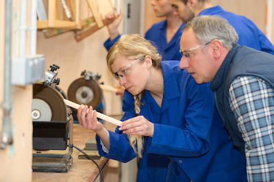 supervisor watching female apprentice carpenter use grinding wheel