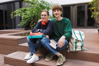 Couple of smiling students sitting with folders and books in hands and happily looking in camera in courtyard of university